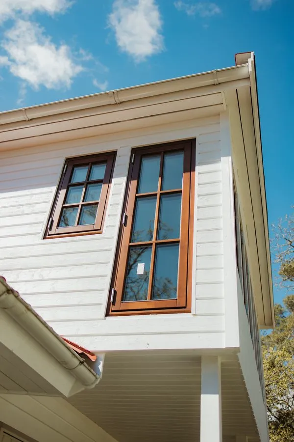 Warm-toned timber windows on a white timber-clad house under blue sky, made by Windows Latvia